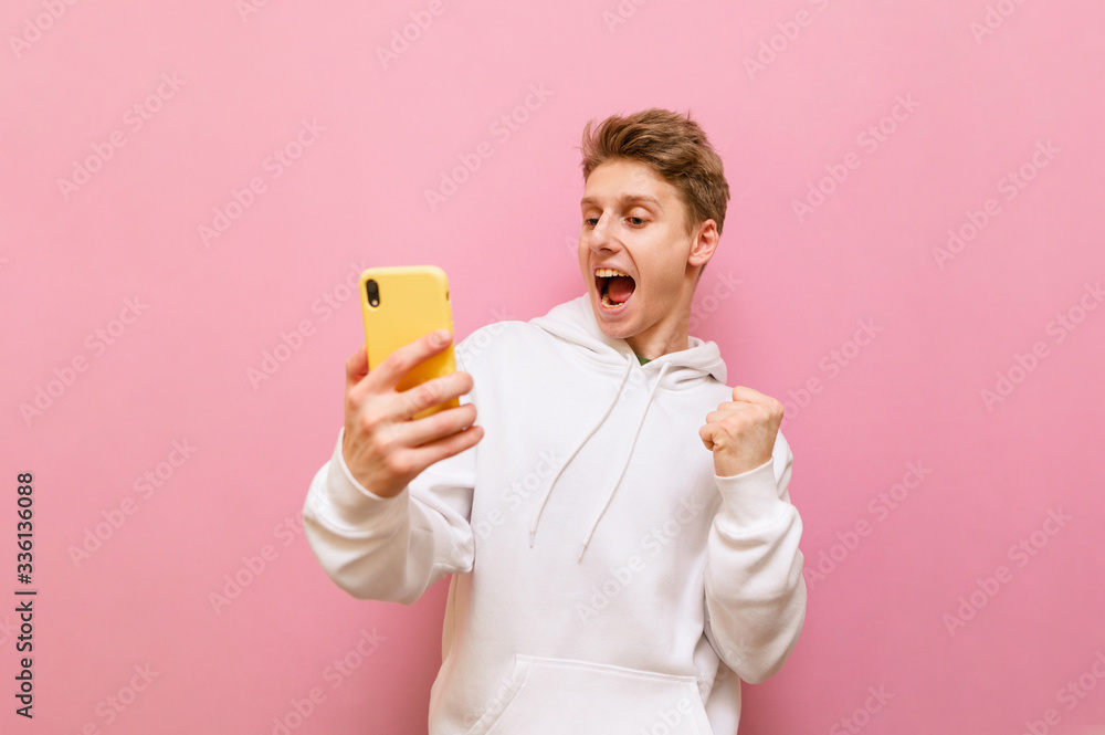 Joyful young man stands on a pink background with a smartphone in his hand, looks at the screen with a happy face and rejoices. The guy is happy to win. Joyful young man stands on a pink background with a smartphone in his hand, looks at the screen with a happy face and rejoices. The guy is happy to win.
