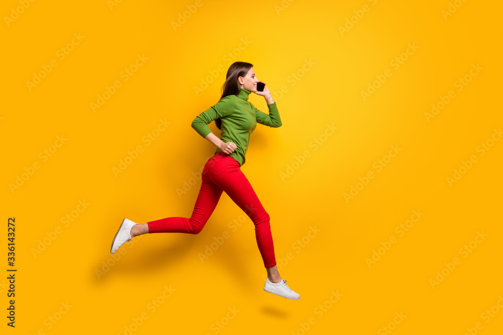 Full length profile photo of funny lady jumping high crazy rushing to meet friends speaking over telephone wear casual green jumper red pants isolated yellow color background Full length profile photo of funny lady jumping high crazy rushing to meet friends speaking over telephone wear casual green jumper red pants isolated yellow color background