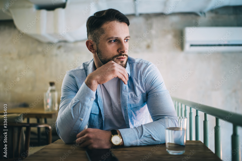 Dreamy caucasian male in trendy shirt and with wristwatch looking away thinking about plans and ideas on break in coffee shop, brunetta 20s hipster guy sitting at table in cafeteria concentrated