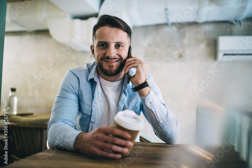 Half length portrait of smiling hipster guy holding disposable cup with beverage talking on mobile phone, cheerful bearded male looking at camera having smartphone conversation on coffee break