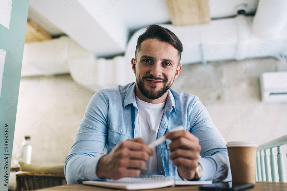 Fototapeta premium Smiling bearded male student satisfied with learning and writing hometask in cafe interior looking at camera holding pen, portrait of cheerful hipster guy sitting at coffee shop thinking about project