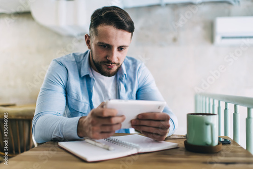 Serious caucasian man millennial holding touchpad playing online game passing next levels, concentrated hipster girl student sitting at cafe interior watching webinar for online course using tablet