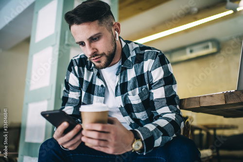 Pensive 20s male millennial man using mobile phone and earbuds for watching movie on free time during coffee break, serious hipster guy reading publication in influencer blog via smartphone in cafe