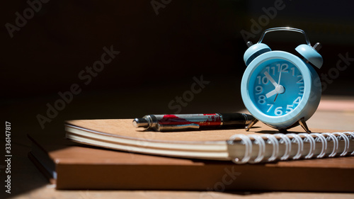 Sunlight and shadow on surface of little blue vintage alarm clock with ballpoint pen and notebooks on wooden table in black background, close up with copy space