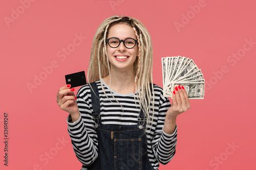 Excited teenager with dreadlocks in a striped shirt and jeans jumpsuit holding cash and bank card.