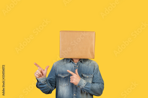 Hombre con caja de cartón en la cabeza con brazos levantados y manos con dedos apuntando hacia la derecha sobre fondo amarillo liso brillante aislado. Vista de frente. Copy space