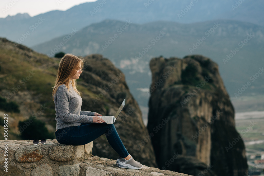 Young female tourist works remotely at laptop at Meteora mountains.
