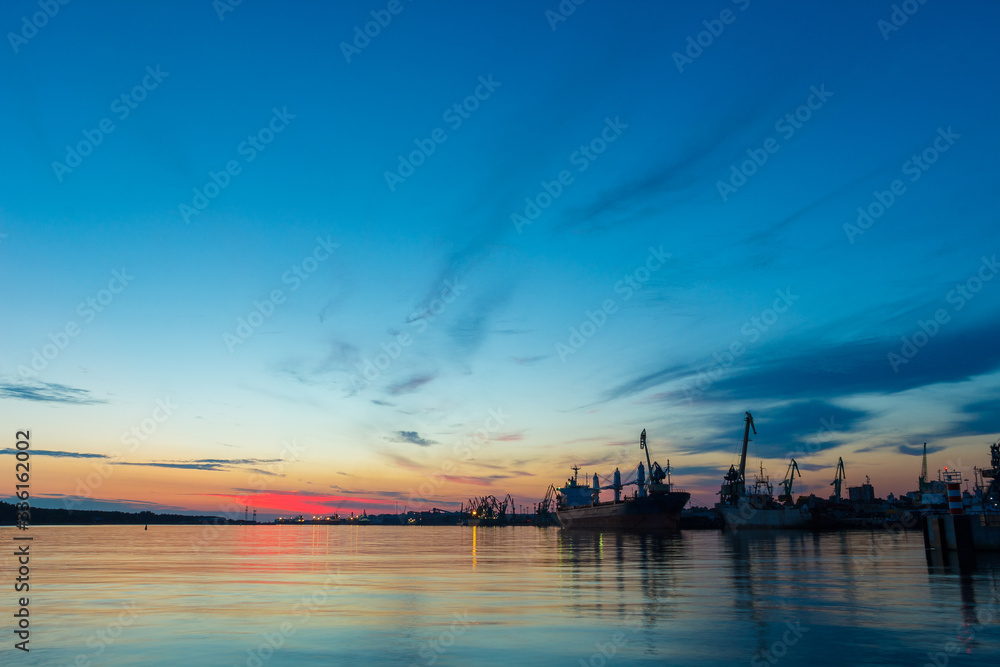 Evening scenery of bay when sun sets. Old harbor with silhouette of sailboats, ships, cranes. Blue and red sky. Nobody. Seaside evening in Klaipeda. Walking along the sea pier. Night scenery in port.