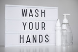 coronavirus pandemic and hand hygiene concept - light box with wash your hands message and bottles of sanitizer or liquid soap over white