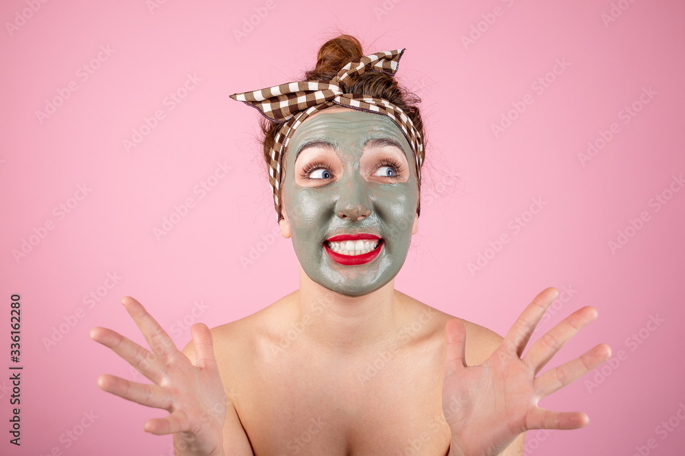 Portrait of a girl in a clay mask on her face standing on a pink background, scared by the burning skin of the mask. Satisfied girl on a skin care procedure Portrait of a girl in a clay mask on her face standing on a pink background, scared by the burning skin of the mask. Satisfied girl on a skin care procedure