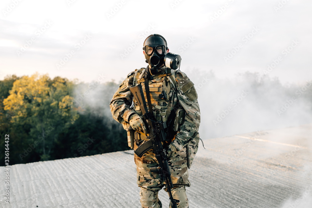 Armed soldier in a respirator with a military machine gun in his hands, standing on the roof of a building on the street against a smoky cloud