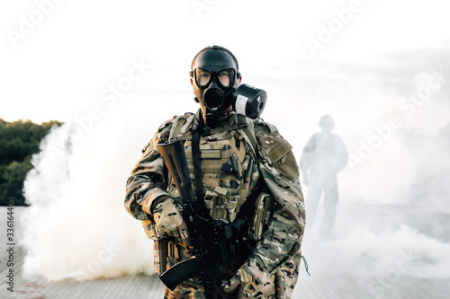 Armed soldier in a gas mask with a rifle standing on the roof of a building in smoke. Strona protection from disorder associated with the coronavirus pandemic