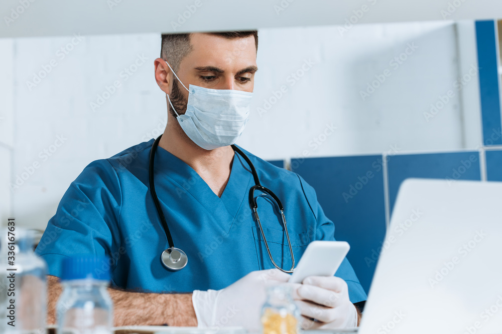 young doctor in medical mask and latex gloves using smartphone near laptop young doctor in medical mask and latex gloves using smartphone near laptop