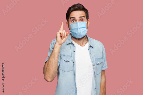 I have an idea. Portrait of surprised young man with surgical medical mask in blue shirt standing with amazed face  looking at camera with idea sign. indoor studio shot  isolated on pink background
