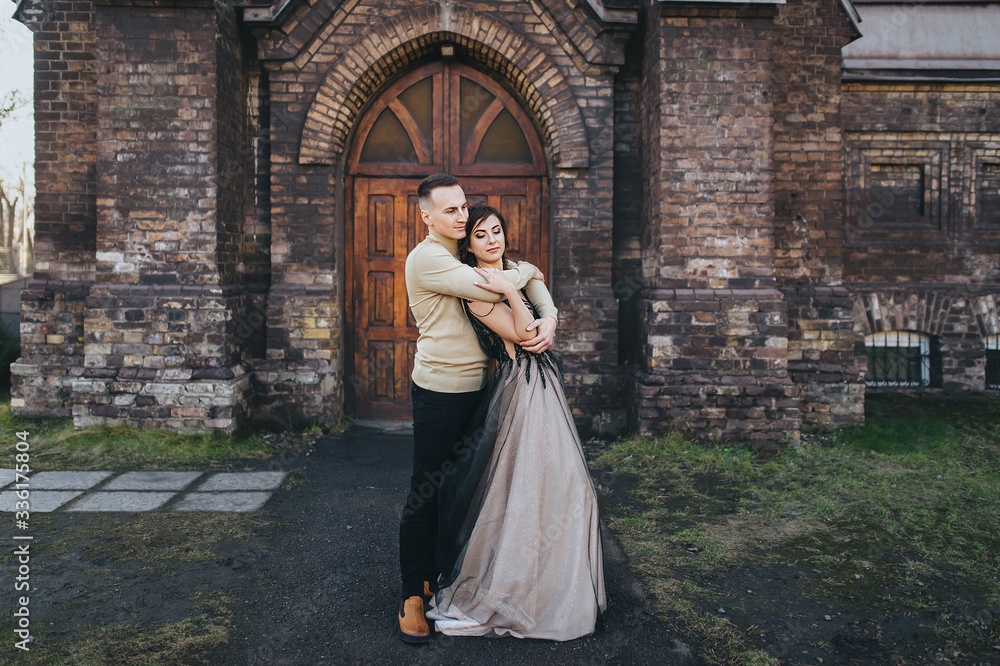 Stylish groom and sweet bride in an expensive dress are hugging against the background of an old church. Wedding portrait of lovers newlyweds. Tender photography, concept.