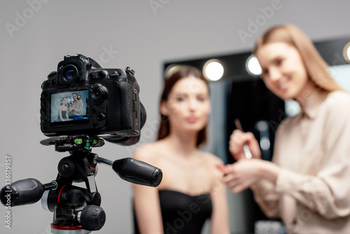 selective focus of digital camera with cheerful makeup artist holding lip gloss near model on screen isolated on grey