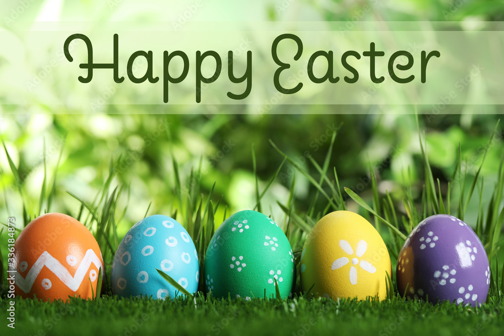 Colorful eggs in green grass and text Happy Easter on blurred background