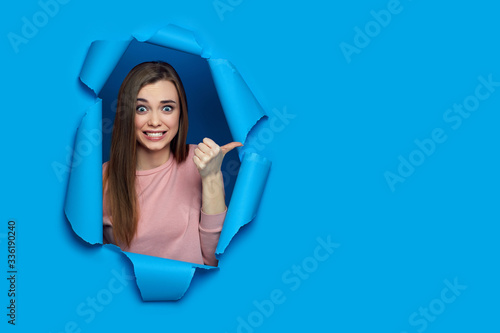 Portrait of beautiful smiling caucasian woman pointing on empty space for your advertising and looking at the camera in torn paper blue wall. banner for discounts and sales © producer