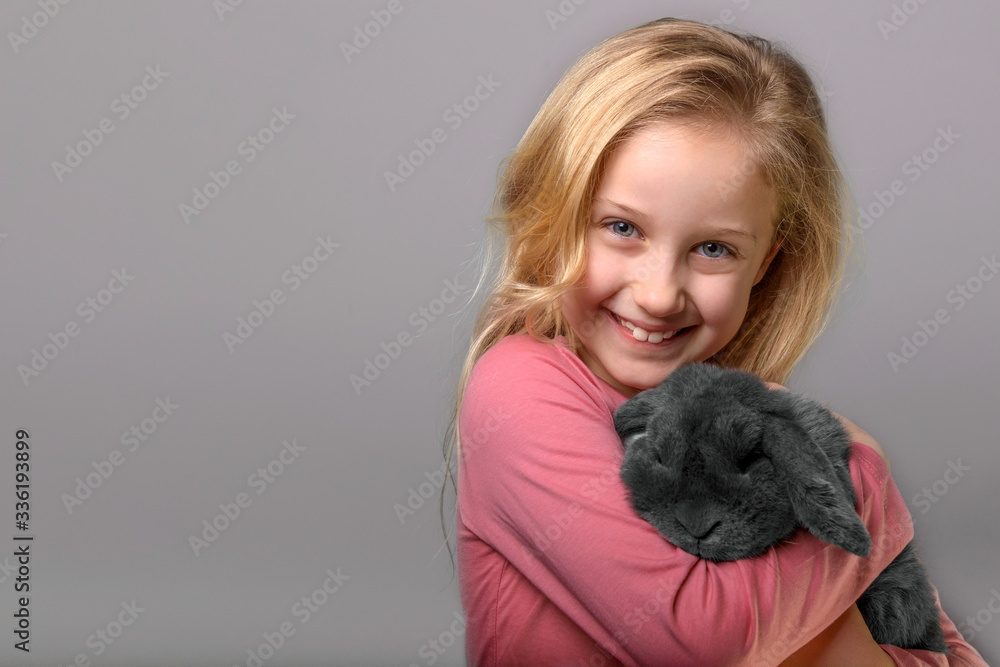 portrait of smiling little girl with rabbit posing in studio, kid girl is holding a little grey rabbit on the grey background, studio shot