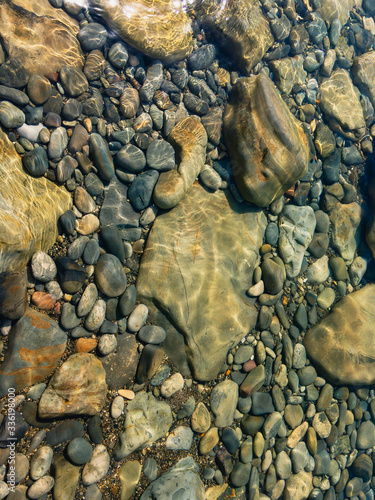 The seabed covered with transparent greenish water with colorful pebbles and sun glare.