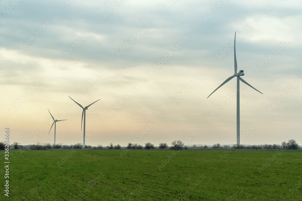 Wind turbines in beautiful agricultural fields under blue sky. Green energy from the windmill park, huge generator during sunset in the meadow. Electric power production technology in the farm. Wind turbines in beautiful agricultural fields under blue sky. Green energy from the windmill park, huge generator during sunset in the meadow. Electric power production technology in the farm.