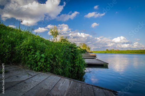 Wide angle view of a waterfront wooden pier surrounded by green vegetation, under a blue sky with puffy clouds