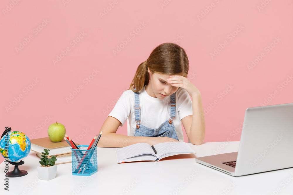 Displeased tired little kid schoolgirl 12-13 years old study at white desk with pc laptop isolated on pink background. School distance education at home during quarantine concept. Put hands on head. Displeased tired little kid schoolgirl 12-13 years old study at white desk with pc laptop isolated on pink background. School distance education at home during quarantine concept. Put hands on head.