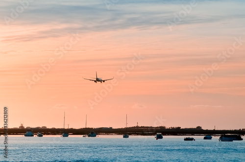 The plane flies to the airport over the river in the city of Faro in Portugal. silhouettes of airplanes and airport, boats in the river, evening city... © SValeriia