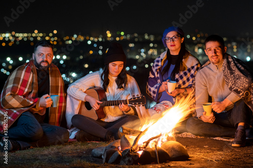 Group of people having fun sitting near bonfire outdoors at night playing guitar, singing songs and talking happily together.