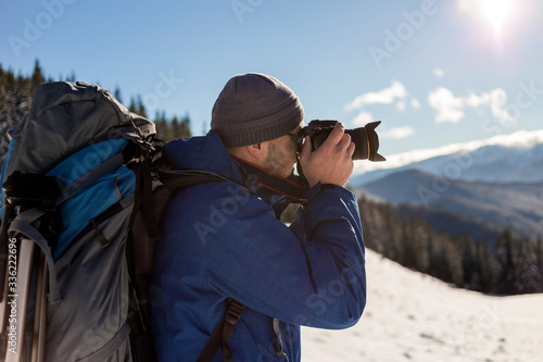Wallpaper Mural Hiker man tourist photographer in warm clothing with backpack and camera taking picture of snowy valley and woody mountain peaks landscape under blue sky on sunny winter cold day. Torontodigital.ca