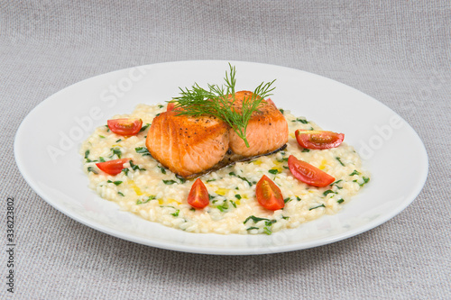 A large piece of fried salmon with rice porridge and butter, decorated with greens and tomatoes, as well as dill on a large round plate on a gray background
