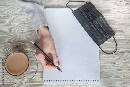 Hand holds a pencil over a notebook. Nearby is a cup of coffee and a black medical mask. Work from home. Stay  home in the coronavirus. Pandemic concept. photo