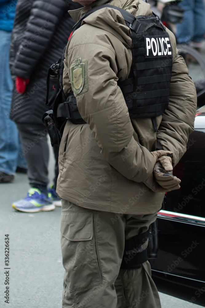 Fototapeta premium A male police officer wearing a khaki parka down-filled uniform and a black bulletproof vest with the word police on the back. There are people in the distance about 6 feet away from the officer.