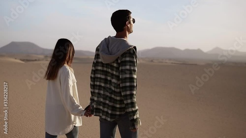 Happy couple holding hands walking in romantic relationship under sun and blue sky in desert. Two young lovers walking by desert sand in casual clothes and smiling photo