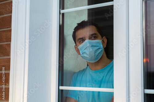 Isolation at home for self quarantine. Young man in medical mask is looking out window. Guy stay home because of quarantine to prevent spreading of coronavirus. Coronavirus pandemic. Social distancing