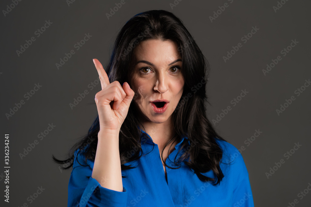 Portrait of adult thinking woman having idea moment pointing finger up on grey studio background. Smiling happy lady showing eureka gesture Portrait of adult thinking woman having idea moment pointing finger up on grey studio background. Smiling happy lady showing eureka gesture