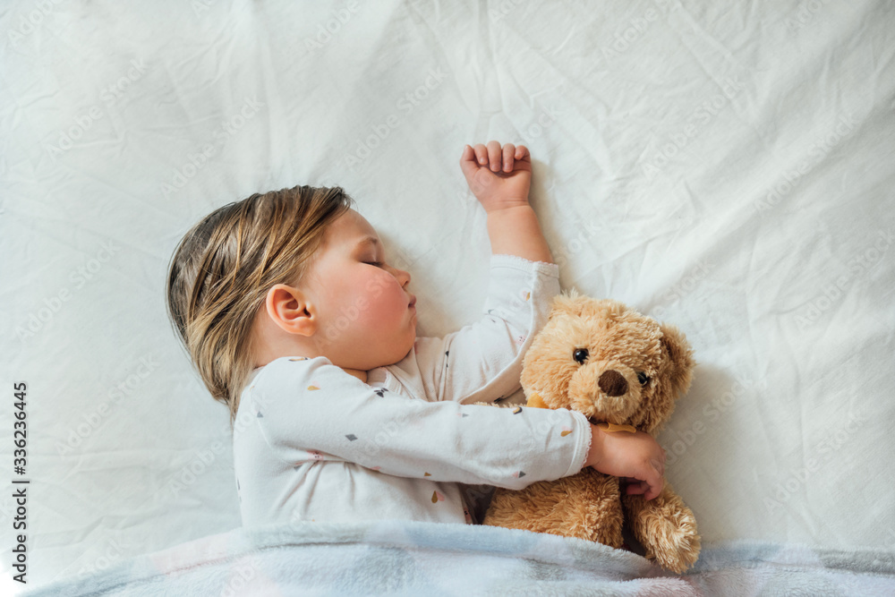 Toddler baby sleeping on white sheets hugging soft brown teddy bear. Children's nap time or bedtime. Child sleep deprivation. Child sleep routine and schedule. Top view. Toddler baby sleeping on white sheets hugging soft brown teddy bear. Children's nap time or bedtime. Child sleep deprivation. Child sleep routine and schedule. Top view.
