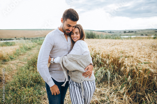 Happy couple in love hugging, kissing and smiling against the sky in field. Hat in girl's hand