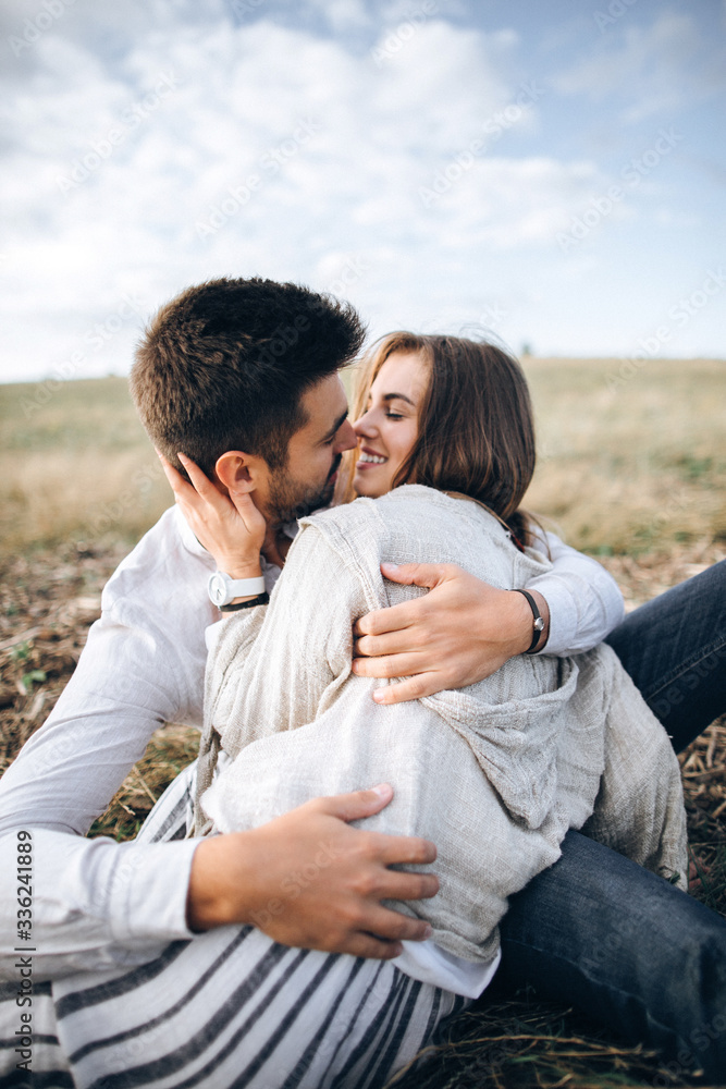 Lovely Сouple hugging, kissing and smiling against the sky seating on grass. Space for text
