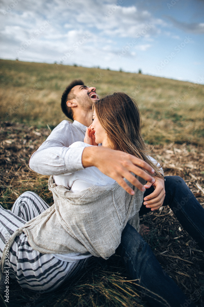 Lovely Сouple hugging, kissing and smiling against the sky seating on grass Lovely Сouple hugging, kissing and smiling against the sky seating on grass