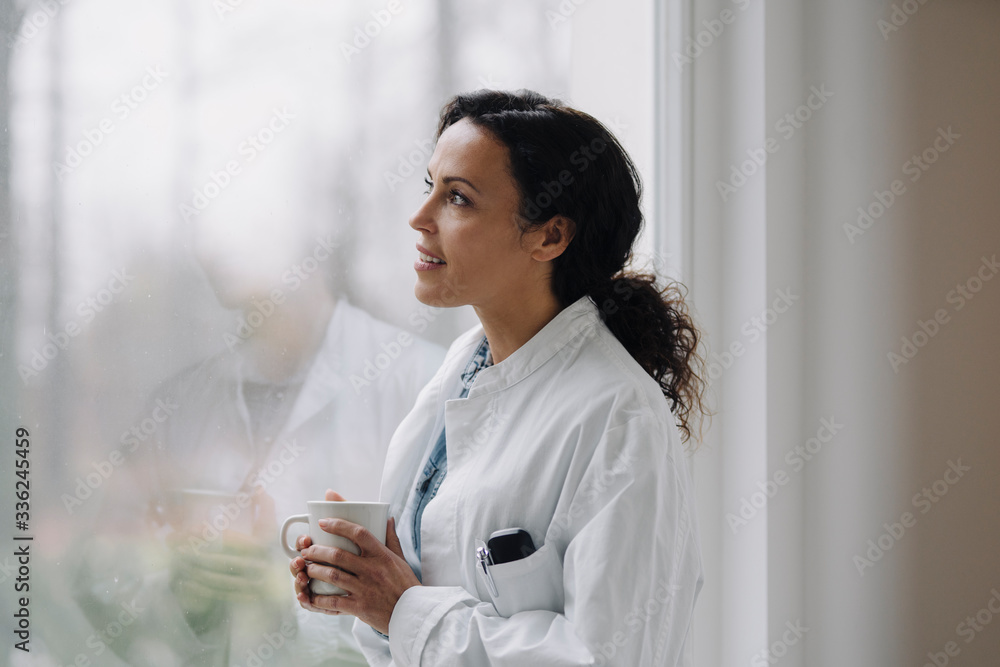 Female doctor looking out of window, holding cup of coffee Female doctor looking out of window, holding cup of coffee