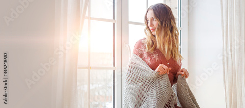 Thoughtful blonde european female wearing beige dress sits on a windowsill and looks outside