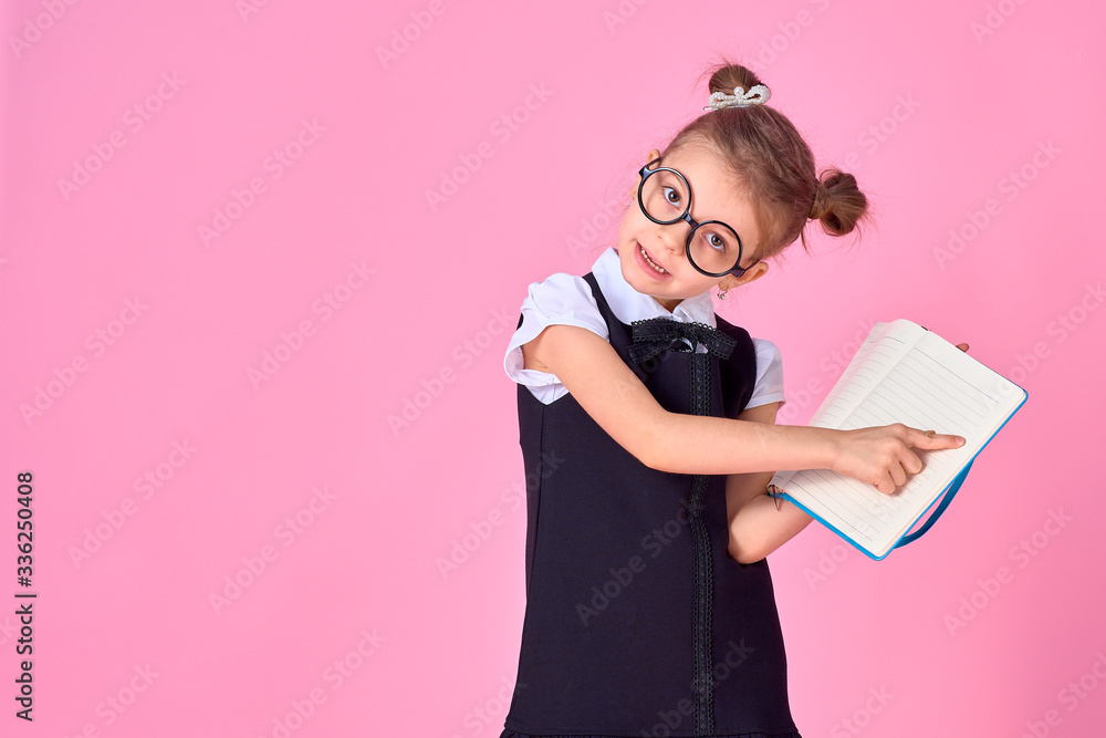 primary school girl in uniform, round glasses without lenses holds a notebook in her hands and points her finger at a blank page on a pink background in the studio. Isolate