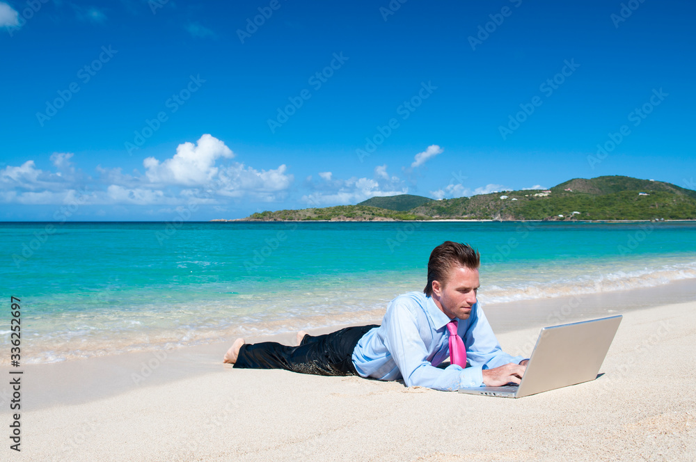 Solitary businessman working remotely on his laptop computer lying outdoors on the shore of a bright tropical island beach Solitary businessman working remotely on his laptop computer lying outdoors on the shore of a bright tropical island beach