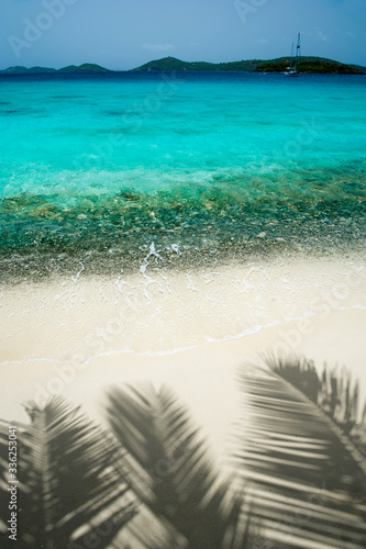 Shadows of palm fronds line the white sand of a vibrant blue tropical island beach