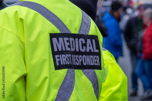 A bright yellow medical first responder uniform being worn by a large male in a crowded street.The coat has a grey reflective cross across the back of the emt or ent. There are people in the distance.