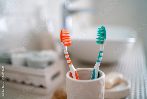 Two colorful toothbrushes stand in a ceramic glass against the background of the washbasin and other accessories in the bathroom