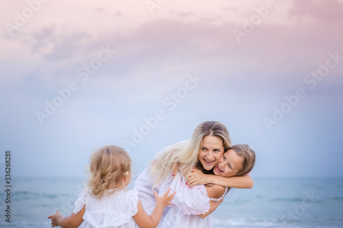 Mom with daughters blondes in white dresses laugh and hug near the blue sea on the beach at sunset. © Alla