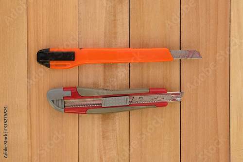 Two stationery knives in orange and red on a wooden background. Office supplies top view. Old stationery knives with narrow blades.