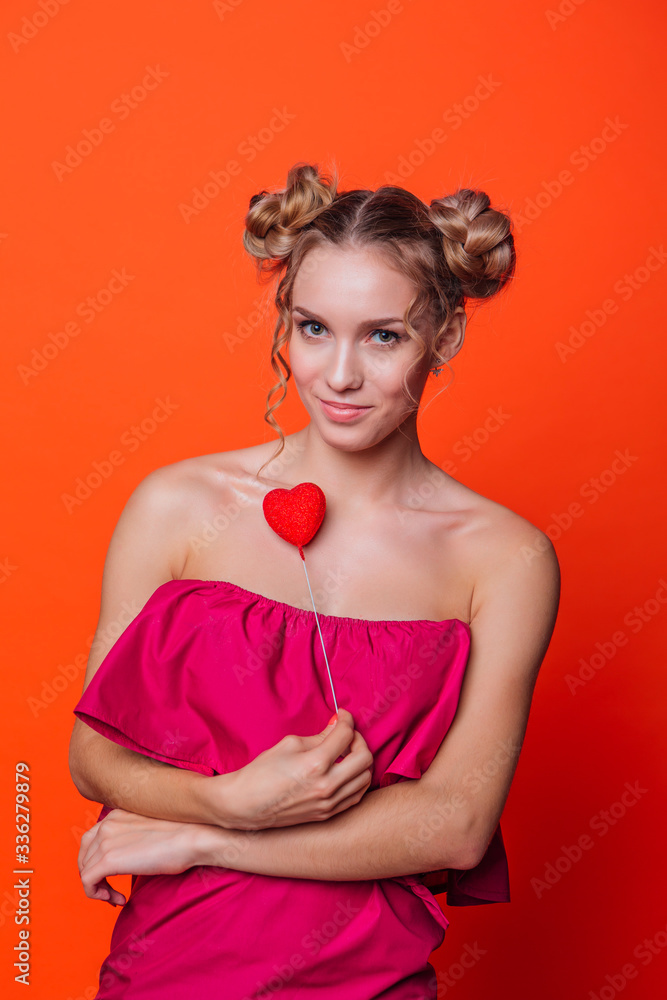 The girl in a red dress on a orange background in the studio. Blonde girl with two hair knots holding red heart on the stick, looking to the camera and smiling. The girl in a red dress on a orange background in the studio. Blonde girl with two hair knots holding red heart on the stick, looking to the camera and smiling.